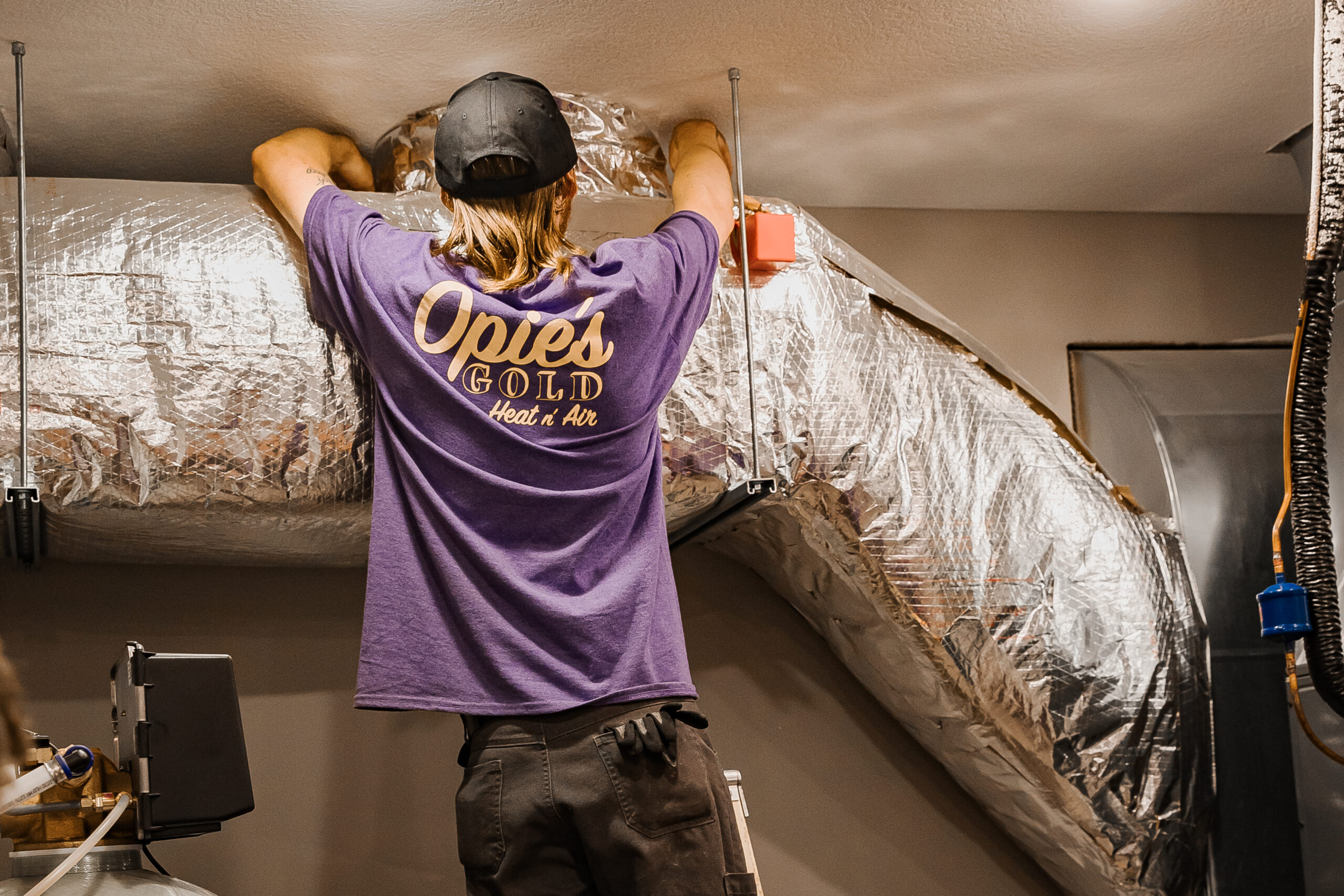 A person in a purple T-shirt and cap adjusts the ductwork in the ceiling.