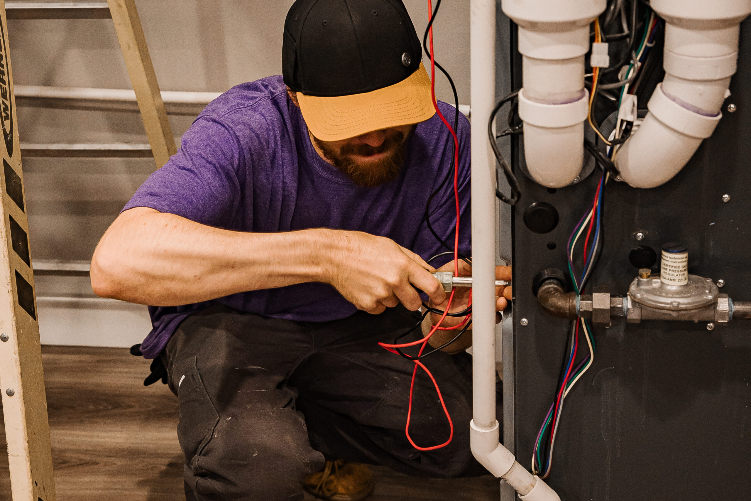 A man wearing a black and orange cap repairs wiring and pipes in a mechanical system using tools.