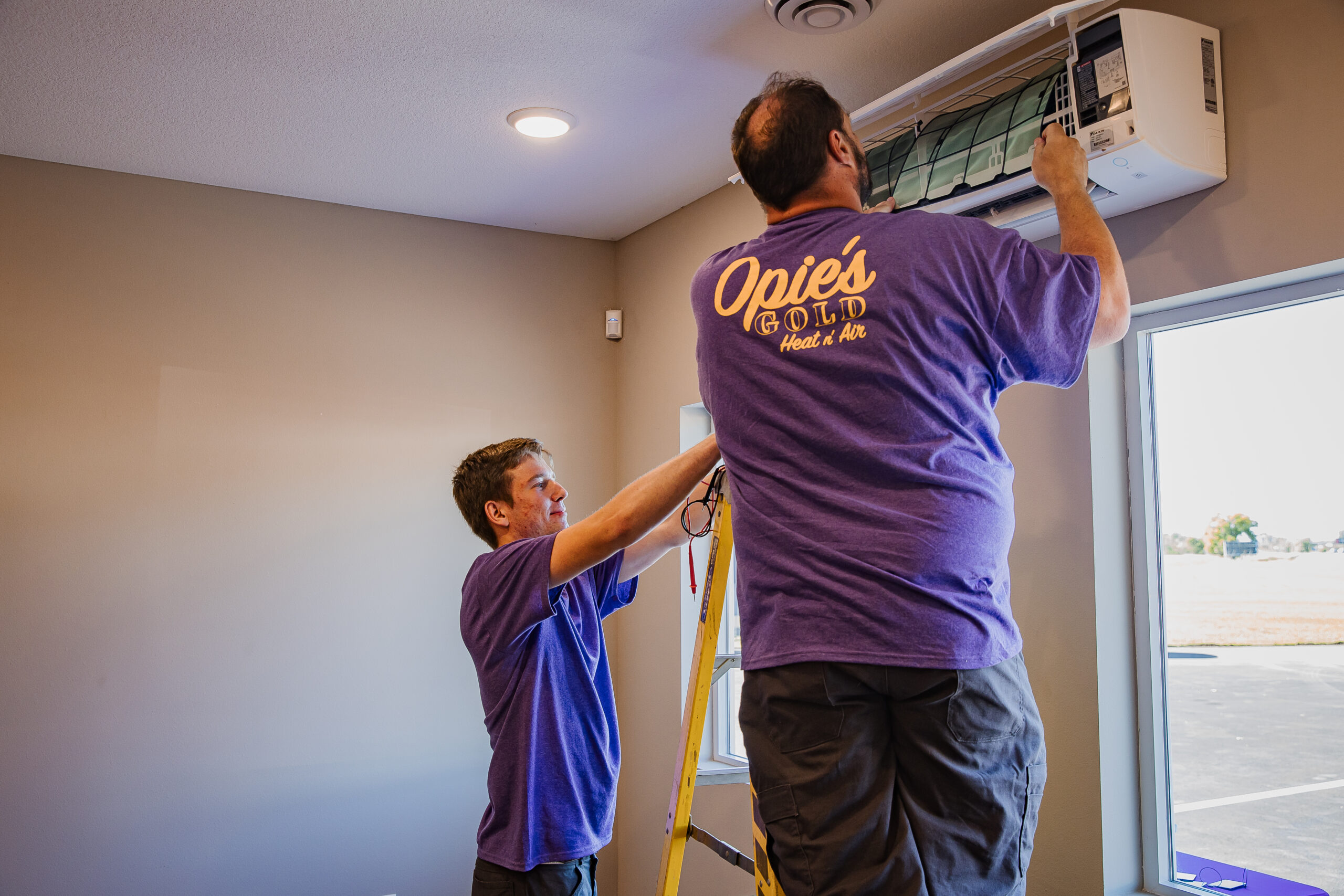 Two technicians in purple shirts work on an air conditioning unit. One climbs a ladder to adjust the device while the other assists inside a room with beige walls.