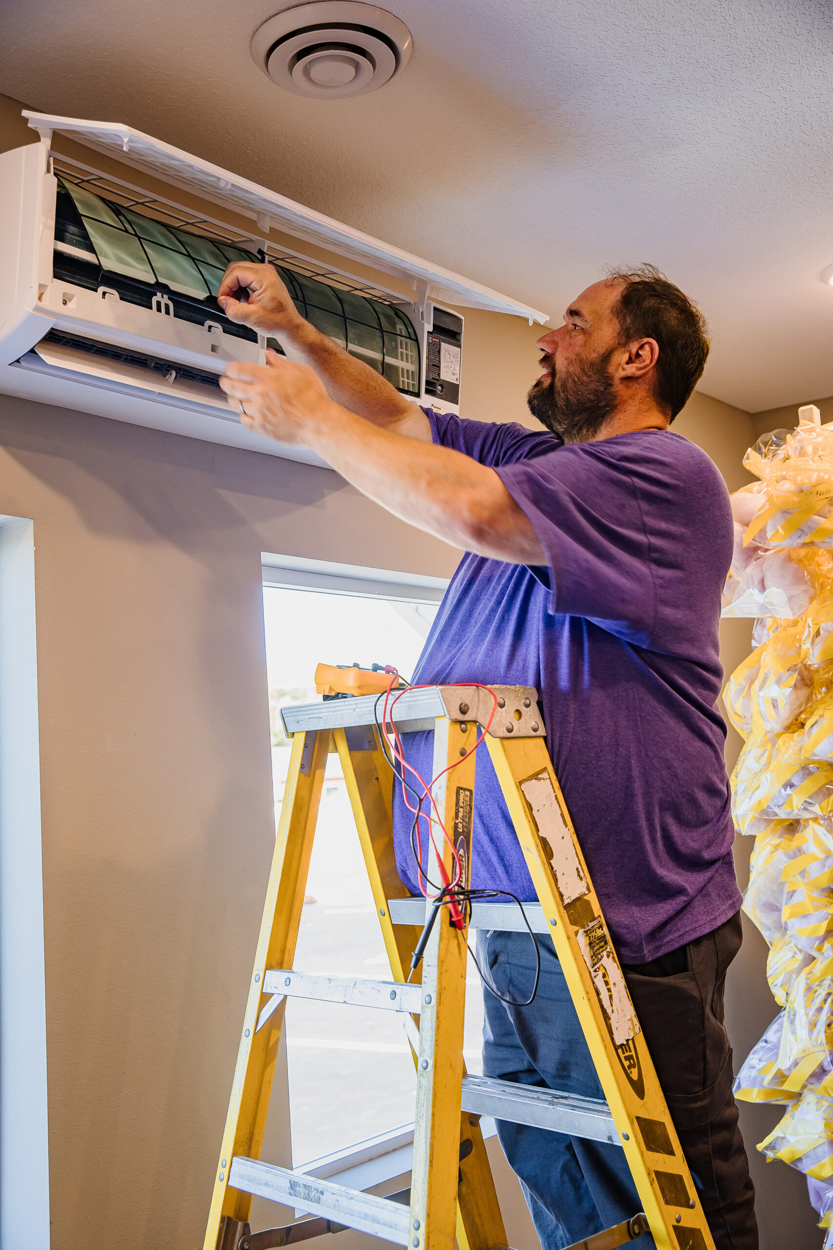 A man repairs a wall-mounted air conditioning unit near a window, surrounded by tools and wires as he stands on a ladder.
