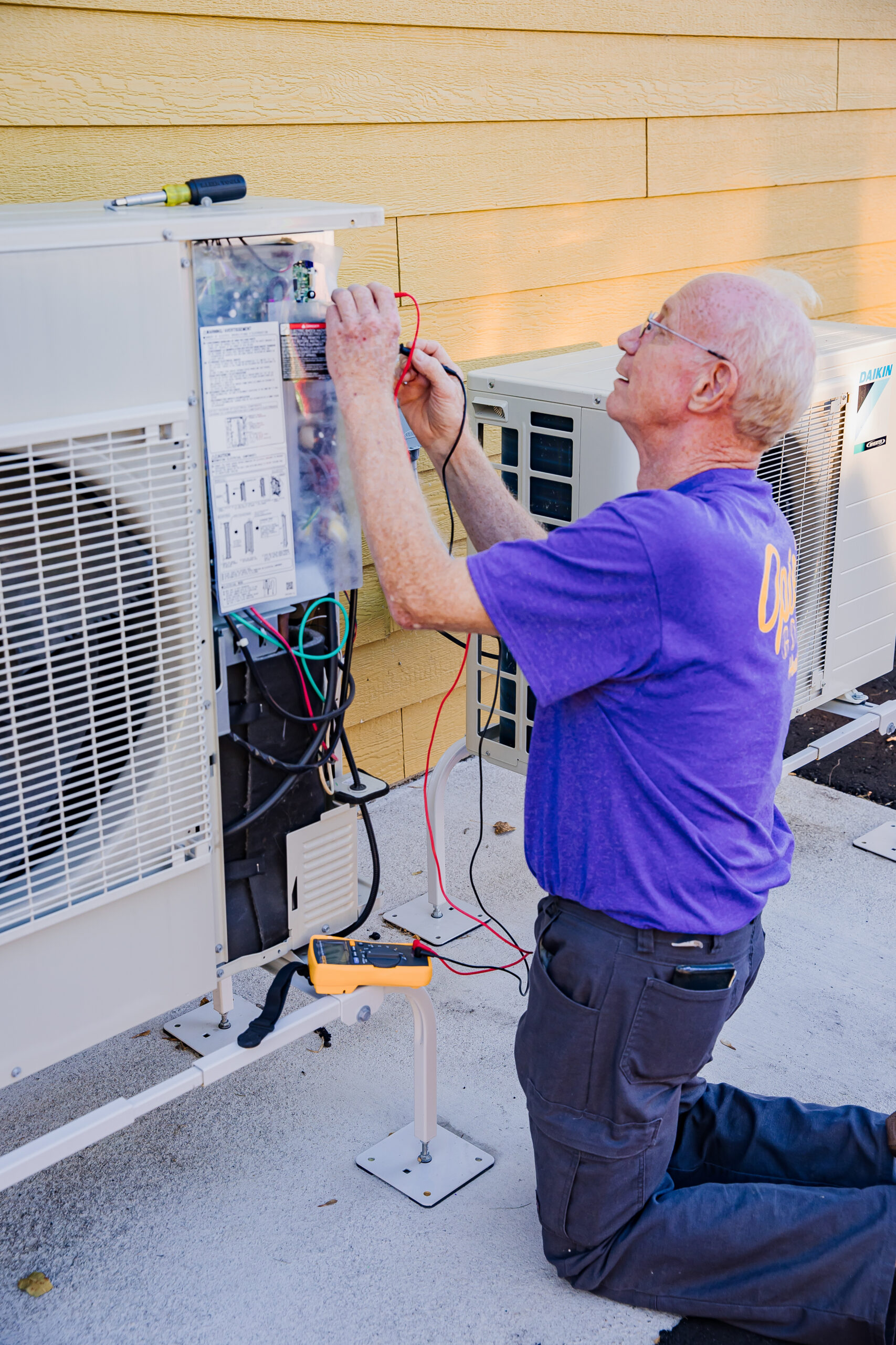 An Opie's Gold Heat n' Air technician kneels and examines an outdoor air conditioning unit.